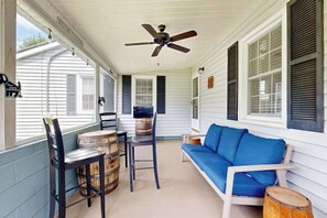 Eine überdachte Veranda mit Deckenventilator, einem blauen Sofa, Holzstühlen und einem Tisch.