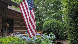 A wooden cabin with an American flag, surrounded by lush greenery and hydrangea flowers. - Rocky Knob Getaway (Scaly Mountain)