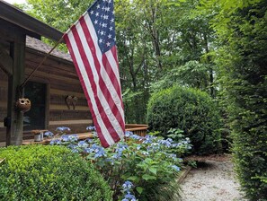 A wooden cabin with an American flag, surrounded by lush greenery and hydrangea flowers. - Rocky Knob Getaway (Scaly Mountain)