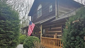 A wooden cabin with a porch, an American flag, and dense greenery. - Rocky Knob Getaway (Scaly Mountain)