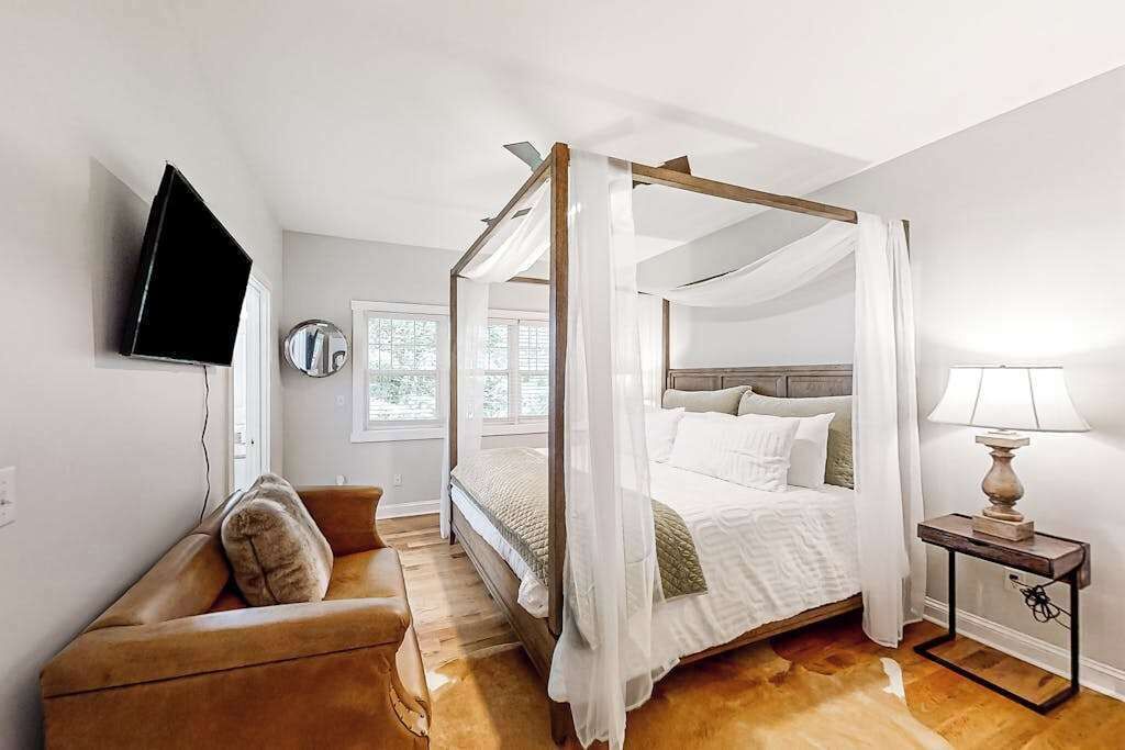 A four-poster bed with a canopy, a television mounted on the wall, a bedside table with a lamp, and a leather sofa.