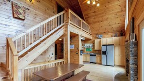 Interno di una baita in legno con una scala in legno, un tavolo da pranzo con panche, un angolo cottura e un camino in pietra.