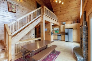 A wooden cabin interior with a wooden staircase, a dining table with benches, a kitchen area, and a stone fireplace. - Bear Bottoms Welcome (Whittier)