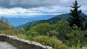 Un banco de piedra con vistas a un paisaje montañoso, con abundante vegetación y cielo despejado.