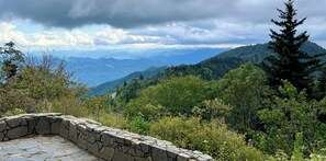 Eine Steinsitzbank mit Blick auf eine gebirgige Landschaft, dichtes Grün und einen klaren Himmel.