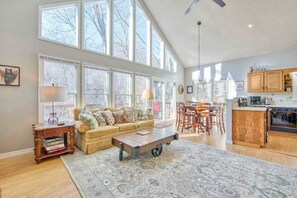 A spacious living area with a sofa, coffee table, and dining table, large windows, and a kitchen area. - Cozy home with wraparound deck, fireplace, AC, & W/D (Waynesville)