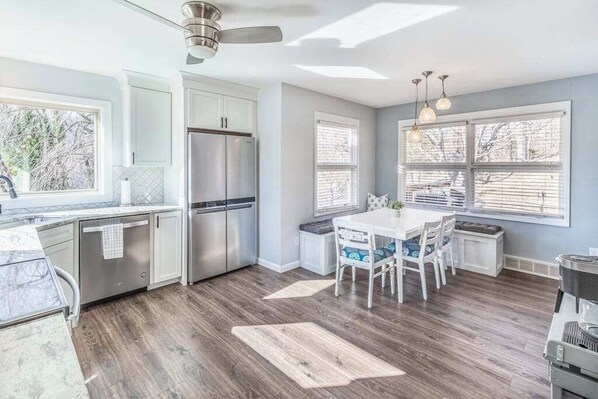 A modern kitchen with white cabinets, a stainless steel refrigerator, and a dining area with a white table and chairs. - The Willows (Waynesville)