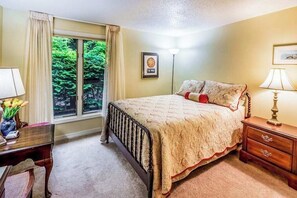 Ein Schlafzimmer mit großem Fenster, einem Bett mit gemustertem Überwurf, einem Nachttisch mit Lampe und einem Schreibtisch mit Blumen.'