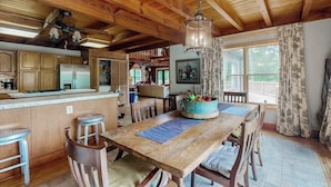 A rustic kitchen with wooden beams, a central island, bar stools, and a dining table with a fruit bowl. - Walela Cabin (Topton)
