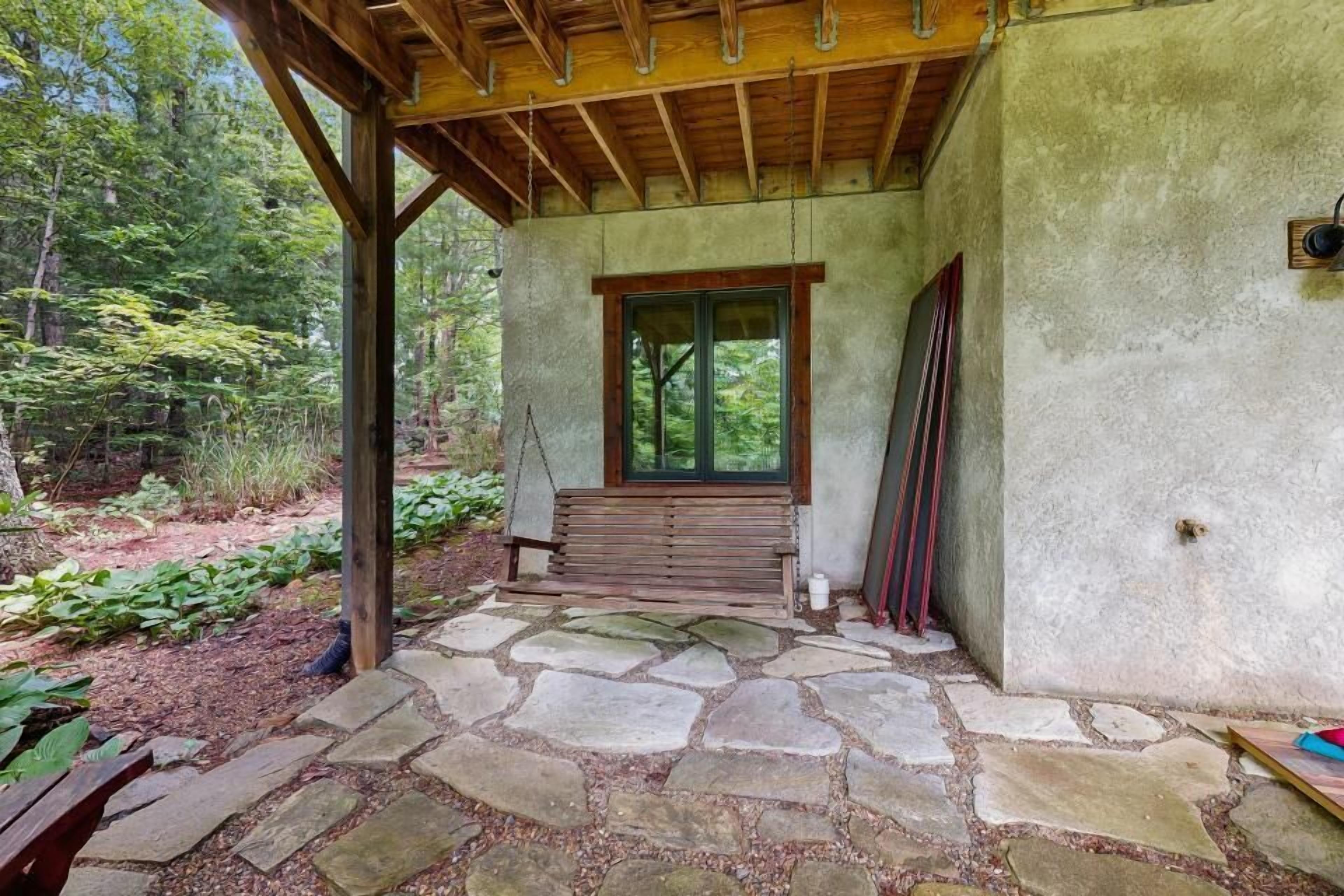 A stone-paved patio with a wooden bench, a window, and a wall-mounted light fixture.