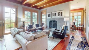A spacious living room with a fireplace, wooden ceiling beams, large windows, and a blue patterned rug. - Eagles Wing Lodge | Game Room, Firepit, Dazzling Sunset Views | Near Downtown (Asheville)