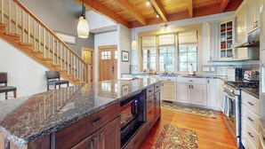 A spacious kitchen with a granite countertop, wooden staircase, and white cabinetry. - Eagles Wing Lodge | Game Room, Firepit, Dazzling Sunset Views | Near Downtown (Asheville)