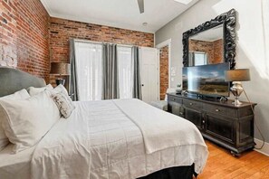 Ein Schlafzimmer mit Backsteinwand, einem Bett mit weißer Bettwäsche, einem Schminktisch, einem Spiegel und einem Fernseher.