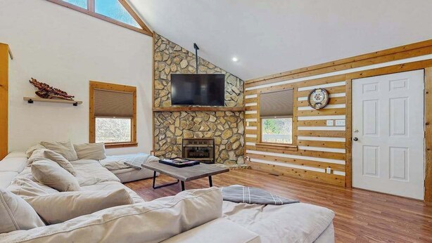 A living room with a stone fireplace, a flat-screen TV, a sofa, and a wooden wall paneling. - Pebble on the Creek (Whittier)