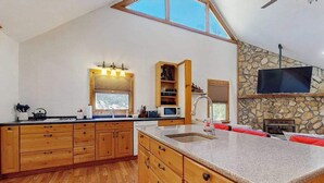 A kitchen with wooden cabinets, a granite countertop, a stone fireplace, and a television mounted on the wall. - Pebble on the Creek (Whittier)