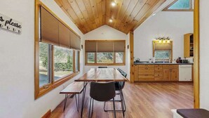 A modern kitchen with wooden cabinets, a dining area with a wooden table and chairs, and large windows with blinds. - Pebble on the Creek (Whittier)