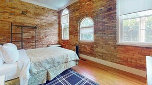 Ein Zimmer mit freiliegenden Backsteinwänden, einem Bett mit gemustertem Überwurf, einem Heizlüfter und einem Fenster mit Blick ins Grüne.