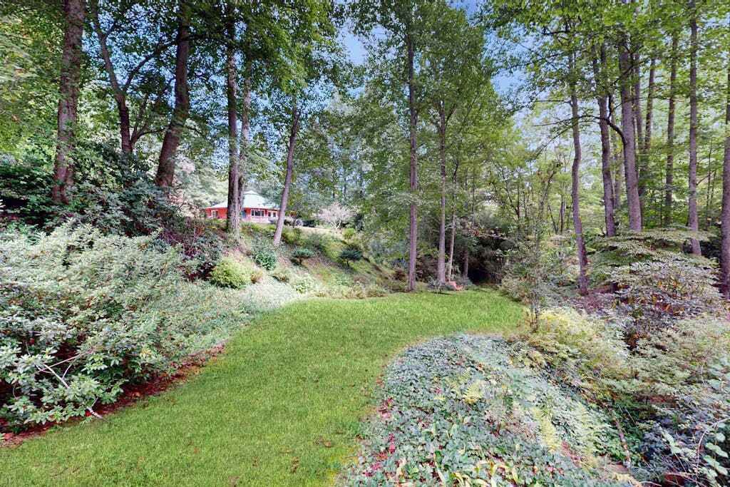 Un jardín con diversas plantas y árboles, un área verde y un edificio rojo al fondo.