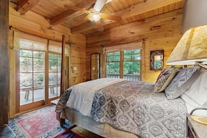 Ein Schlafzimmer in einer Holzhütte mit einem Bett, einem Deckenventilator und großen Fenstern.