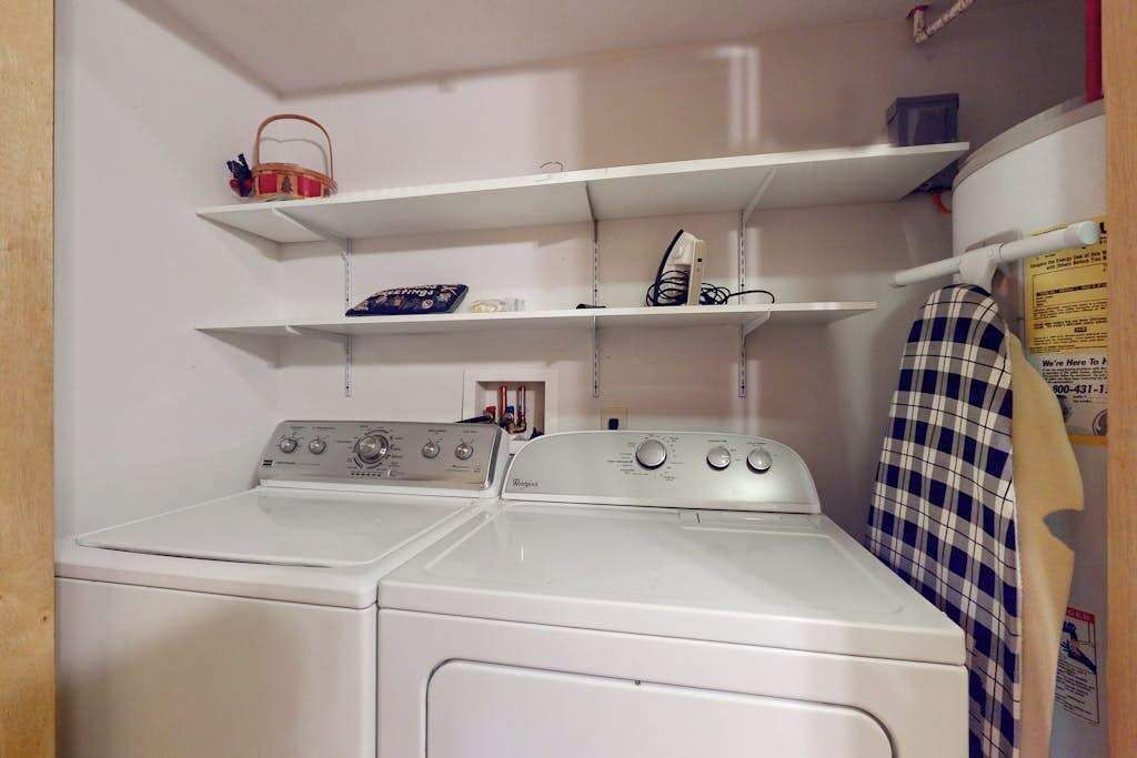 A laundry area with two white washers and dryers, shelves above holding various items, and a checkered towel hanging on the wall.