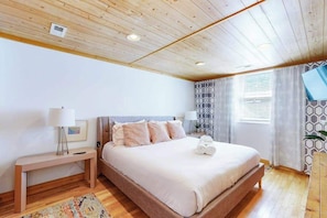 A bedroom with a wooden ceiling, a bed with white bedding, a bedside table with a lamp, and a window with curtains.