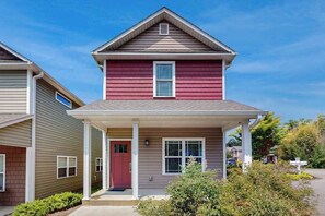 Ein zweistöckiges Haus mit roter Haustür, einer Veranda und einem kleinen Vorgarten mit einem Kreuz.