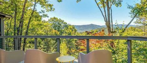 A deck with chairs and a table, overlooking a forested landscape with mountains in the distance.