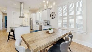 A modern kitchen with white cabinets, a wooden dining table, and a large window with blinds. - Downtown Delight | Luxury home, walk to Main Street, bike to trails (Brevard)