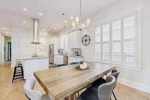 A modern kitchen with white cabinets, a wooden dining table, and a large window with blinds. - Downtown Delight | Luxury home, walk to Main Street, bike to trails (Brevard)