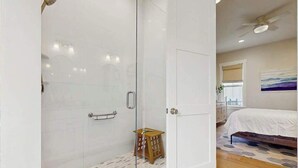 A modern bedroom with a glass shower enclosure, a wooden stool, a bed with a white bedspread, and a ceiling fan. - Downtown Delight | Luxury home, walk to Main Street, bike to trails (Brevard)