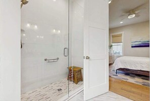 A modern bedroom with a glass shower enclosure, a wooden stool, a bed with a white bedspread, and a ceiling fan. - Downtown Delight | Luxury home, walk to Main Street, bike to trails (Brevard)