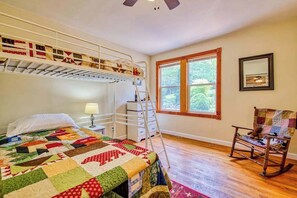 Un dormitorio con literas, colcha, mecedora, escalera, ventana y un cuadro en la pared.