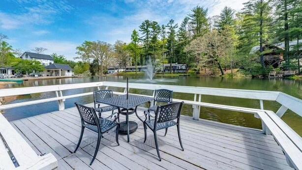 A wooden deck with a table and chairs, overlooking a body of water with trees and buildings in the background. - Lakeview pet friendly studio with pool, fire pit, canoes & paddle boats, & WiFi (Flat Rock)