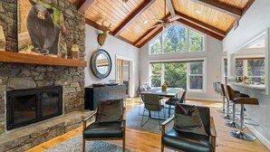 Une pièce spacieuse avec une cheminée en pierre, des poutres apparentes en bois au plafond, un coin repas et de grandes fenêtres.