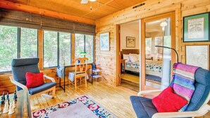 Ein gemütliches Blockhaus-Interieur mit Holzbalkendecke, Deckenventilator, Essbereich mit Stühlen, einem Bett und einem großen Fenster.