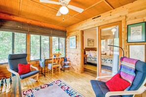 Ein gemütliches Blockhaus-Interieur mit Holzbalkendecke, Deckenventilator, Essbereich mit Stühlen, einem Bett und einem großen Fenster.