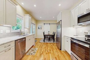 A modern kitchen with white cabinets, stainless steel appliances, and a dining area with a wooden table and chairs. - All-new cottage with deck, grill, & washer/dryer - near downtown & Rail Trail (Hendersonville)