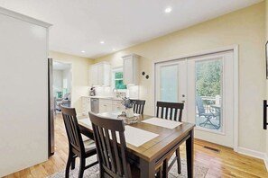 A dining area with a wooden table and chairs, a kitchen in the background, and a sliding glass door leading to a balcony. - All-new cottage with deck, grill, & washer/dryer - near downtown & Rail Trail (Hendersonville)