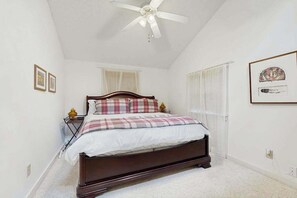 Ein Schlafzimmer mit Bett, Deckenventilator, Nachttisch, Fenster mit Vorhängen und gerahmten Bildern an der Wand.