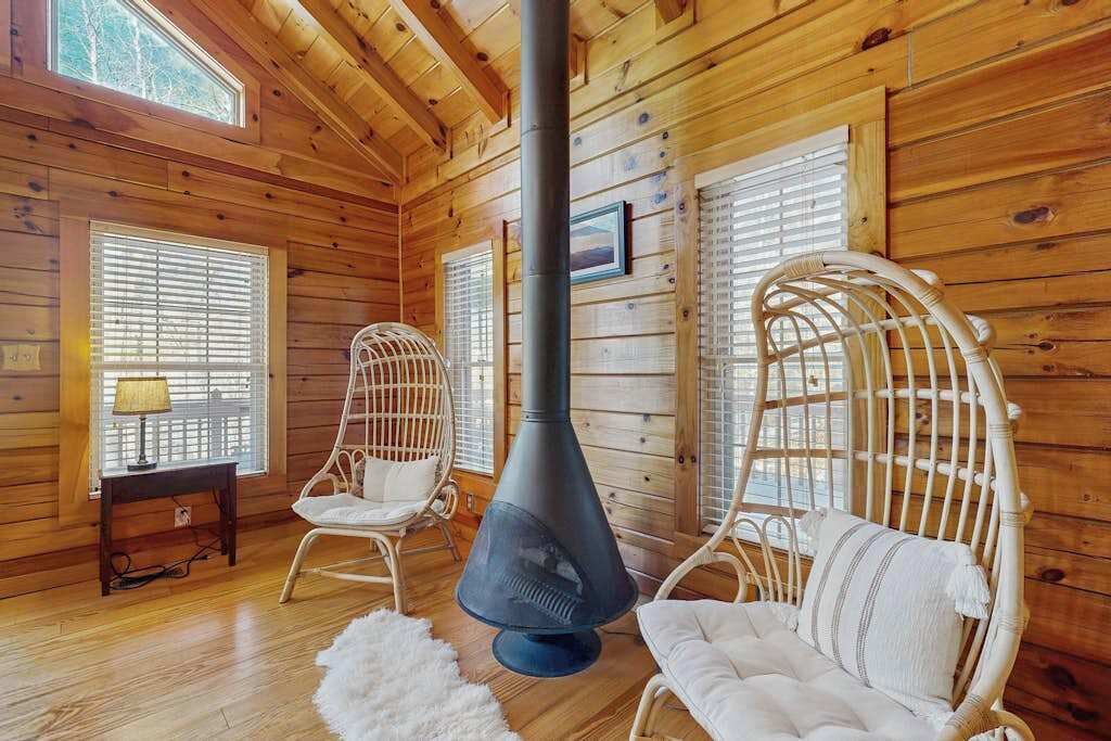 A log cabin interior with wooden walls, two wicker chairs, a black heater, and a window with blinds.