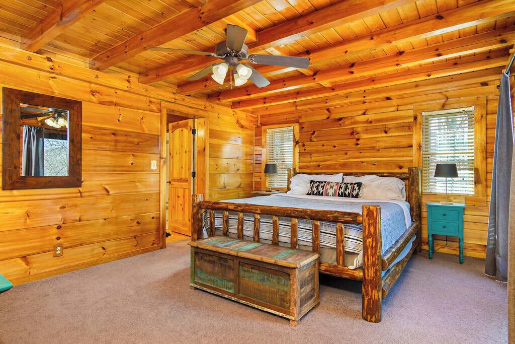 Een slaapkamer in een blokhut met een houten bed, een plafondventilator, een kleine tafel en een bank.
