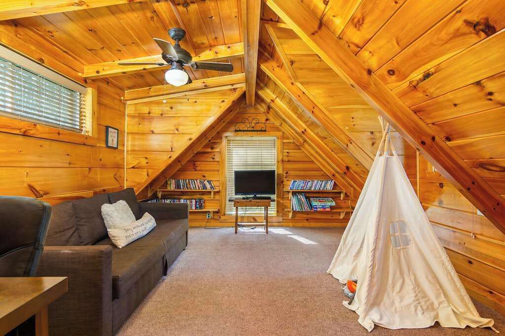 A cozy attic room with a wooden ceiling, a sofa, a TV, a bookshelf, and a tent-like structure.