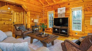 Interior de una cabaña de troncos con techo de madera, televisor, sofá, mesa de centro y ventana.