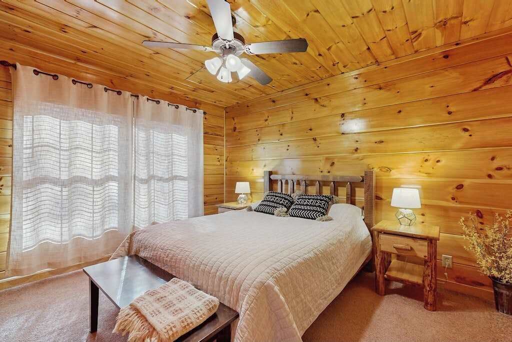 Une pièce aux murs lambrissés, comprenant un lit, un ventilateur de plafond, une table de chevet avec une lampe et une fenêtre avec des rideaux.