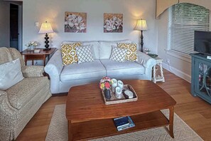 Ein Wohnzimmer mit einer Couch, zwei Beistelltischen mit Lampen, einem Couchtisch mit Dekoration und einem Fernseher auf einer Anrichte.