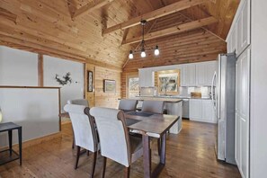 A spacious room with a wooden ceiling, a dining area with chairs, a kitchen with white cabinets, and a refrigerator. - Mountain view home with large deck, private hot tub and fire-pit (Fairview)