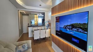 A modern living room with a flat-screen TV mounted on the wall, a dining area with a white table and chairs, and a kitchen in the background.