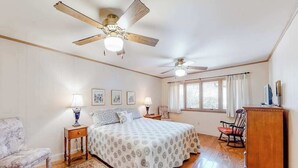 Un dormitorio con una cama, dos mesitas de noche, una silla, un sillón mecedora, un armario de madera, dos ventiladores de techo y una ventana con cortinas.