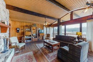 A cozy living room with a fireplace, wooden walls, a large window with a view, and a ceiling fan. - Morris Villa Upper (Lake Junaluska)