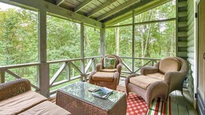 Un porche cubierto con muebles de mimbre, una mesa de vidrio y vistas a un bosque.